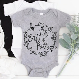 May include: A gray baby onesie with a black floral wreath design and the text "Itty Bitty & Pretty".