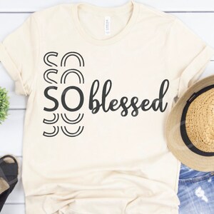 May include: A beige t-shirt with a black graphic that says "SO blessed" with a stylized "SO" design. The shirt is lying on a white wooden surface with a straw hat, blue denim shorts, and black shoes.