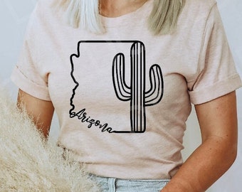 May include: A pink t-shirt with a black outline of the state of Arizona and a cactus inside the outline. The text "Arizona" is written in a cursive font below the outline.