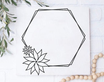 May include: A white square canvas with a black geometric hexagon design and floral accents. The artwork is decorated with a sprig of green leaves and a strand of wooden beads, set against a white brick wall.