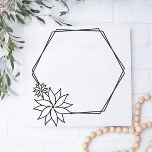 May include: A white square canvas with a black geometric hexagon design and floral accents. The artwork is decorated with a sprig of green leaves and a strand of wooden beads, set against a white brick wall.
