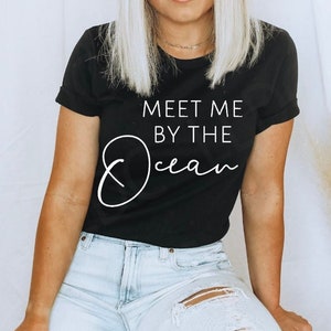 May include: A black t-shirt with white text that reads "Meet me by the Ocean". The model is wearing the shirt with a straw hat and light blue jeans.