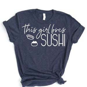 May include: A navy blue t-shirt with the text "This girl loves sushi" in white lettering. The text is accompanied by two white illustrations of sushi rolls.