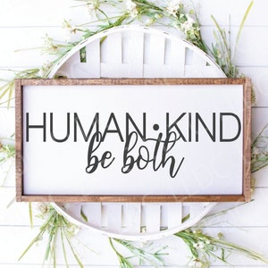 May include: A rectangular wooden sign with the words "HUMAN KIND be both" in black lettering. The sign is surrounded by white flowers and greenery, set against a white background.