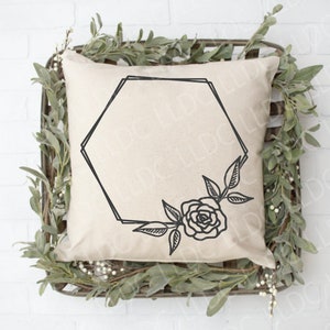Puede incluir: Una almohada blanca con un marco hexagonal negro y un diseño floral con una sola rosa. La almohada está sentada en un lecho de follaje verde.