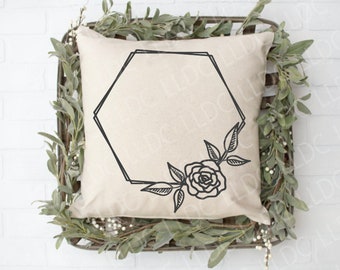 May include: A white pillow with a black hexagonal frame and a floral design featuring a single rose. The pillow is sitting on a bed of green foliage.