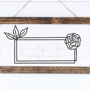 Puede incluir: Una impresión en lienzo blanco con un contorno negro de una rosa y hojas. La impresión tiene un marco negro con el texto "Lovely Life Designs Co." dentro del marco.