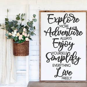 Puede incluir: Un letrero de madera blanco con texto en caligrafía negra que dice "Explore More Adventure Always Enjoy Yourself Simplify Everything Live Freely". El letrero está colgado en una pared de ladrillo blanco.