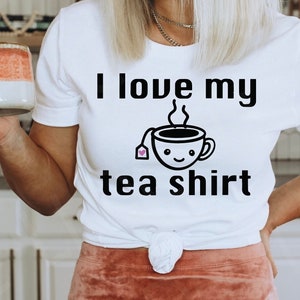 K&ouml;nnte beinhalten: Wei&szlig;es T-Shirt mit schwarzem Text, der "Ich liebe mein Tee-Shirt" lautet. Der Text wird von einem Cartoon-Bild einer Teetasse mit einem Teebeutel und Dampf begleitet.