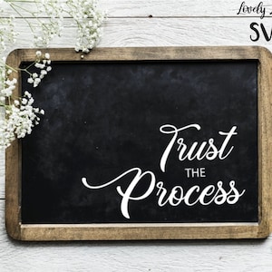 May include: A black chalkboard with a wooden frame, featuring the text "Trust the Process" in white cursive lettering. The chalkboard is on a white wooden surface with white flowers and green leaves.