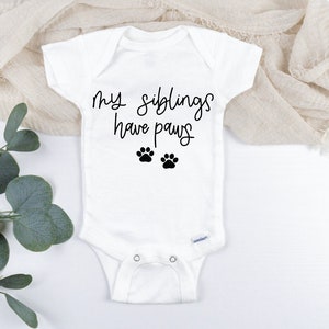 Op de afbeelding: Witte baby romper met zwarte tekst die "Mijn broertjes en zusjes hebben pootjes" zegt en twee zwarte poot afdrukken.