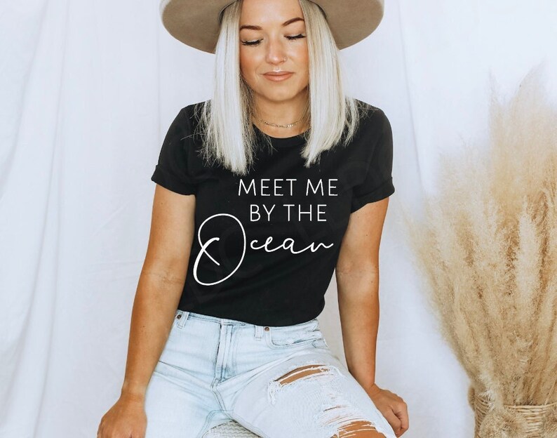 May include: A black t-shirt with white text that reads "Meet me by the Ocean". The model is wearing the shirt with a straw hat and light blue jeans.
