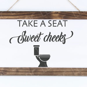 Puede incluir: Un cartel blanco con marco de madera marr&oacute;n y el texto "Take a seat Sweet cheeks" con una silueta negra de un inodoro.