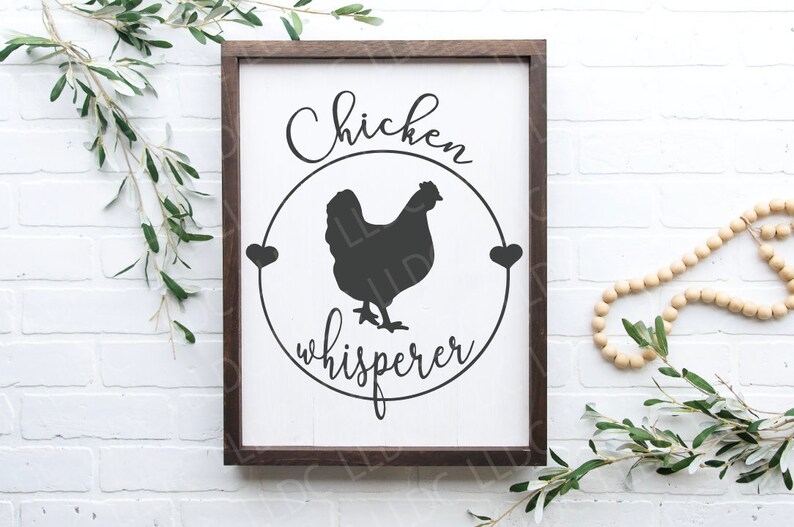 May include: A white wooden sign with a brown frame, featuring a black silhouette of a chicken inside a circle with the text "Chicken whisperer" in black cursive lettering.