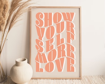 May include: A framed print with the text "SHOW YOURSELF MORE LOVE" in a peachy-pink colour on a beige background. The print is framed in a light wood frame and is sitting on a woven rug.