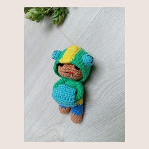 Op de afbeelding: Een handgemaakte gehaakte pop in een groene, gele en blauwe outfit. De pop heeft een capuchon met blauwe oordetails, een groen jasje, een blauwe broek en bruine schoenen. De pop heeft een kleine rode tong.