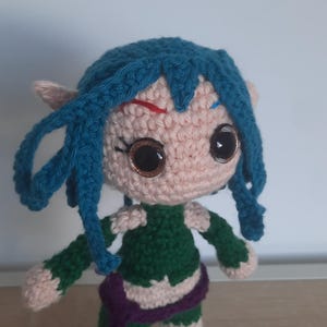 May include: A crocheted doll with blue hair, green clothing, and brown eyes. The doll is standing on a wooden surface.