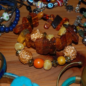 Könnte beinhalten: Eine Sammlung von Armbändern in verschiedenen Stilen und Farben. Das zentrale Armband hat bernsteinfarbene Steine und dekorative Perlen. Weitere Armbänder sind in Blau-, Türkis- und Goldtönen gehalten, vor einem Holzgrund.