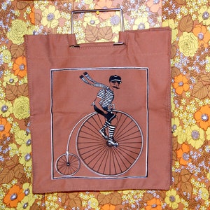 Peut inclure: Sac fourre-tout marron avec une poignée en métal. Le sac présente une illustration encadrée d'un homme à vélo penny-farthing. L'homme porte une chemise rayée, un short et une écharpe. Le fond est un motif floral.