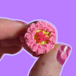 May include: A pink and yellow cake charm with a pink heart design in the center. The charm is made of polymer clay and is about the size of a quarter.