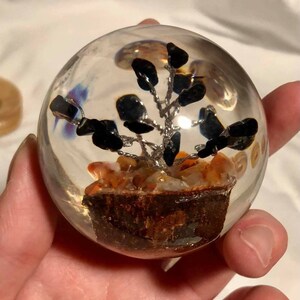 May include: A clear glass sphere with a small tree made of black stones and silver wire inside. The tree is sitting on a bed of orange and brown stones.