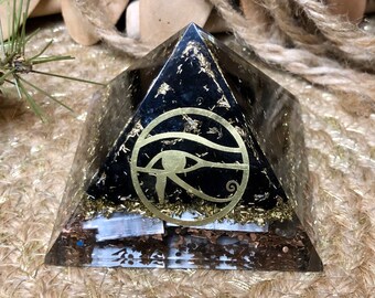 May include: A black pyramid-shaped orgone energy generator with gold flakes and a gold-colored Eye of Horus symbol. The pyramid is set on a textured, neutral-toned surface, creating a contrast with the dark, glossy structure.