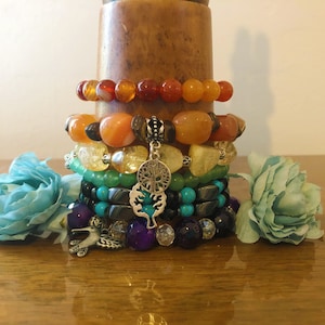 May include: A stack of colorful beaded bracelets with charms, including a hummingbird and a tree of life, on a wooden stand. The bracelets are made of various stones, including carnelian, agate, turquoise, and amethyst.