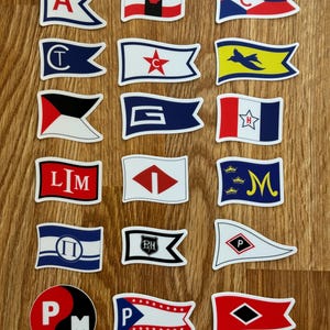 Może przedstawiać: Kolekcja 18 flag sygnałowych morskich z różnymi literami, symbolami i wzorami. Flagi są wydrukowane na białym papierze z czerwonymi, niebieskimi, żółtymi i czarnymi akcentami. Flagi są ułożone w trzy rzędy po sześć flag.