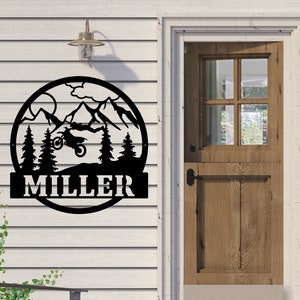 May include: Black metal sign with a silhouette of a dirt bike jumping over mountains. The sign has a circle around the image and the name "MILLER" in a banner below the circle.