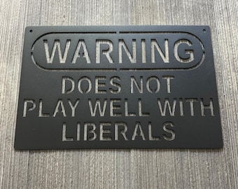 Puede incluir: Letrero de metal negro con las palabras "WARNING DOES NOT PLAY WELL WITH LIBERALS" recortadas. El letrero tiene esquinas redondeadas y dos agujeros para colgar. El letrero se muestra sobre un fondo de madera gris.