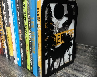 May include: Black metal bookends featuring a silhouette of a moose, trees, and a bird in flight. The bookends are holding up a row of books with titles visible, including "The Underdog Advantage" and "Duck Commander Family".