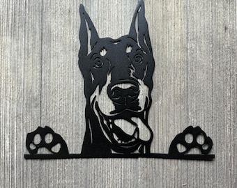 May include: Black metal silhouette of a Doberman Pinscher dog peeking over a sign with its paws on the edge. The dog has a happy expression with its tongue sticking out.
