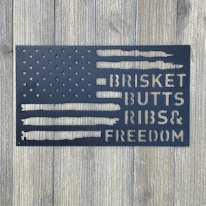 May include: Black metal wall art in the shape of the American flag. The flag design includes cut-out stars and stripes, with the words "BRISKET BUTTS RIBS & FREEDOM" cut into the stripes. The art is mounted on a wooden background.