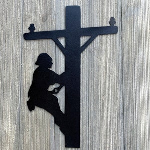 May include: Black metal silhouette wall art depicting a lineman climbing a utility pole. The lineman is wearing a hard hat and is holding onto the pole with one hand. The pole has a crossbar with insulators at the top.