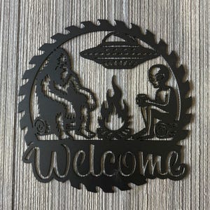 May include: Black metal welcome sign with a silhouette of a Bigfoot and an alien sitting by a campfire with a UFO above them. The sign is shaped like a saw blade.