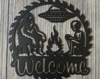 May include: Black metal welcome sign with a silhouette of a Bigfoot and an alien sitting by a campfire with a UFO above them. The sign is shaped like a saw blade.