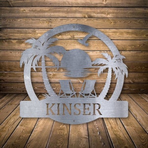 May include: Metal silhouette of a beach scene with two palm trees, a setting sun, and two lounge chairs. The silhouette is in a circle with the name "KINSER" below it.