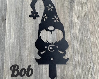 May include: Black metal silhouette of a gnome with a pointed hat and flowers. The gnome is holding a sign that says "Bob".