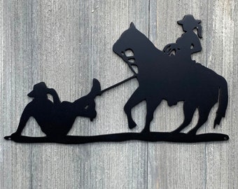 May include: Black metal silhouette wall art depicting a cowgirl on horseback roping a steer. The artwork is mounted on a wood background.