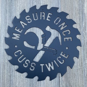 K&ouml;nnte beinhalten: Schwarzes Metallschild in Form einer S&auml;ge mit dem Text "Measure Once, Cuss Twice" und einem Hammer und einer S&auml;ge, die sich in der Mitte kreuzen.