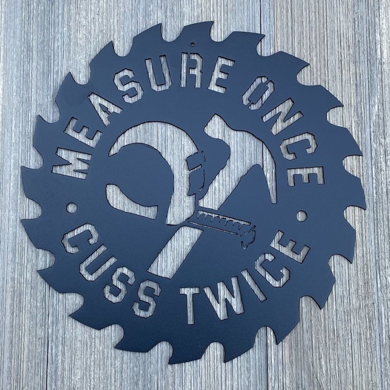 K&ouml;nnte beinhalten: Schwarzes Metallschild in Form einer S&auml;ge mit dem Text "Measure Once, Cuss Twice" und einem Hammer und einer S&auml;ge, die sich in der Mitte kreuzen.