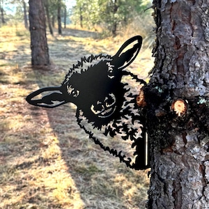 May include: Black metal silhouette of a sheep's head peeking out from behind a tree trunk. The sheep has a fluffy, curly coat and a friendly expression.