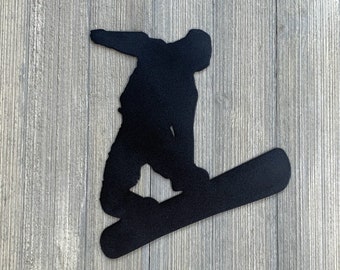 Może przedstawiać: Czarna sylwetka snowboardzisty w powietrzu, wykonującego trik na snowboardzie. Sylwetka jest wycięta z metalu i zamontowana na drewnianym tle.