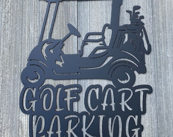 May include: Black metal sign depicting a golf cart with golf clubs and the words "GOLF CART PARKING". The sign is mounted on a wooden background.