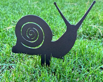 May include: A black metal garden stake in the shape of a snail. The snail has a spiraled shell and two long antennae. The garden art is placed in green grass.