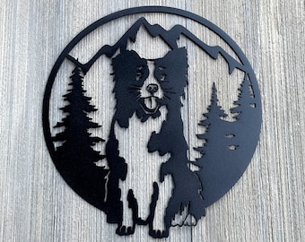 May include: Black metal silhouette of a border collie dog standing in front of a mountain range with pine trees. The silhouette is cut out of a round metal disc.