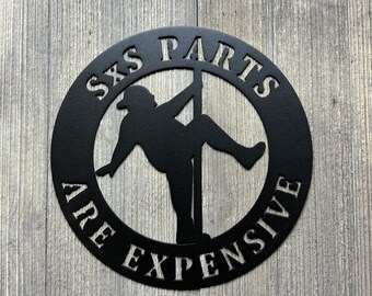 May include: Black metal circular sign featuring a silhouette of a person hanging from a pole. The text around the silhouette reads "SXS PARTS ARE EXPENSIVE".