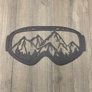 May include: A black metal wall art piece shaped like ski goggles. The goggles feature a cut-out design of a mountain range and evergreen trees. The artwork is mounted on a wooden surface.