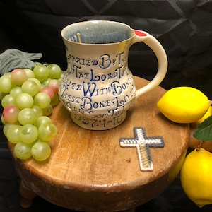 May include: A white ceramic mug with a blue and white design. The mug has a handle and a red dot on the side. The mug is sitting on a wooden surface with a cross, grapes, and lemons.