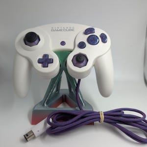 Pode incluir: Um comando Nintendo GameCube branco com botões roxos, um analógico roxo e um stick C roxo. O comando tem um cabo roxo e está pousado num suporte cinzento e vermelho. O logótipo da Nintendo GameCube está impresso no centro superior.